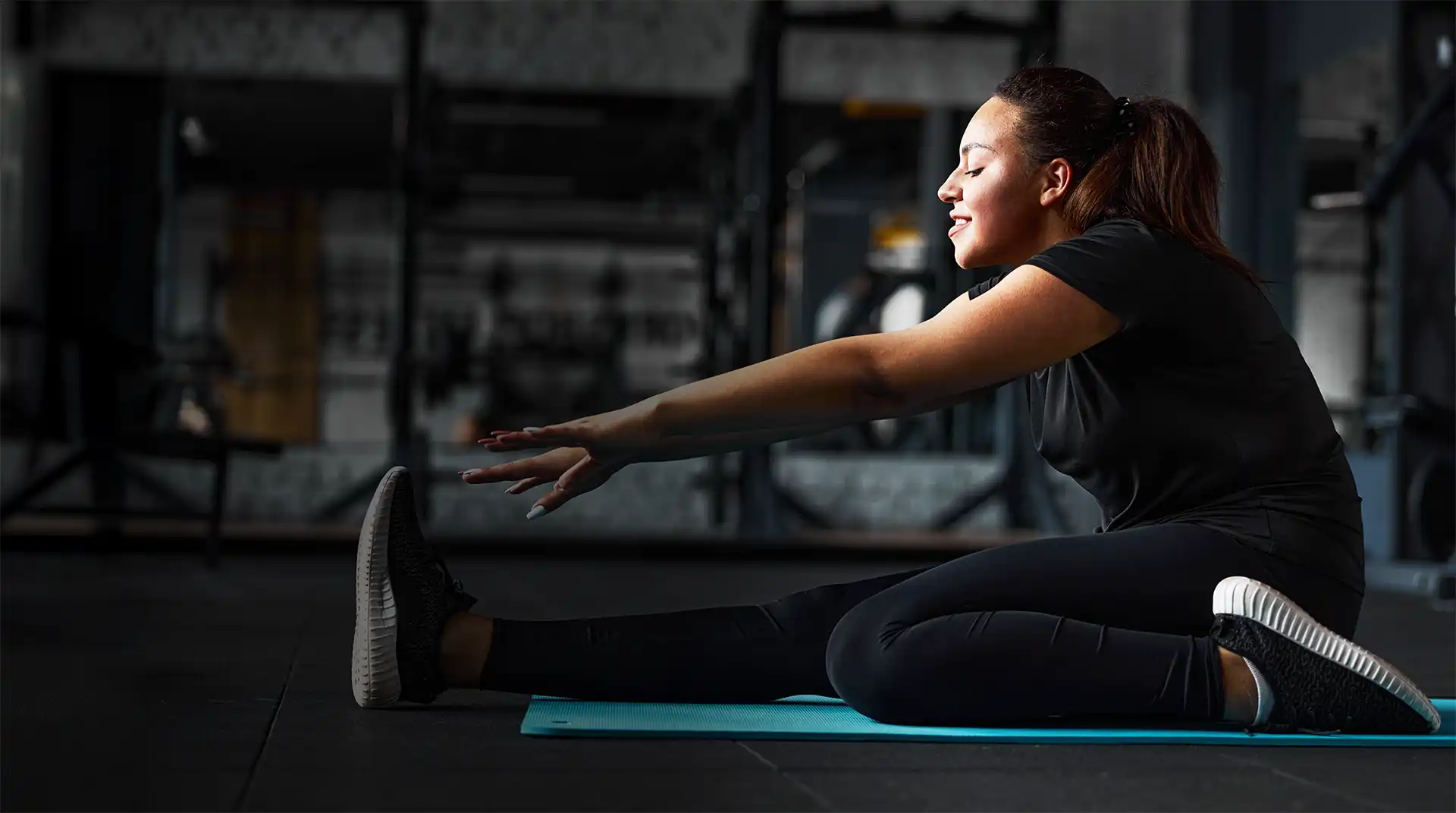 A young woman with a peaceful expression stretches deeply on a blue yoga mat in a modern gym, demonstrating commitment to her physical well-being and personal development goals with nRecovery.