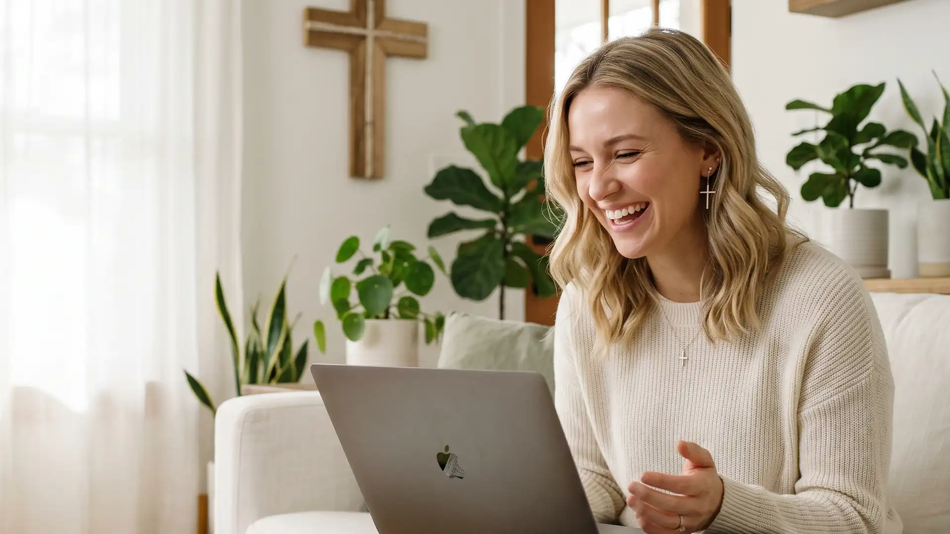 A happy woman with a cross necklace laughing while participating in an online faith-based coaching session on her laptop in a bright, peaceful home environment.