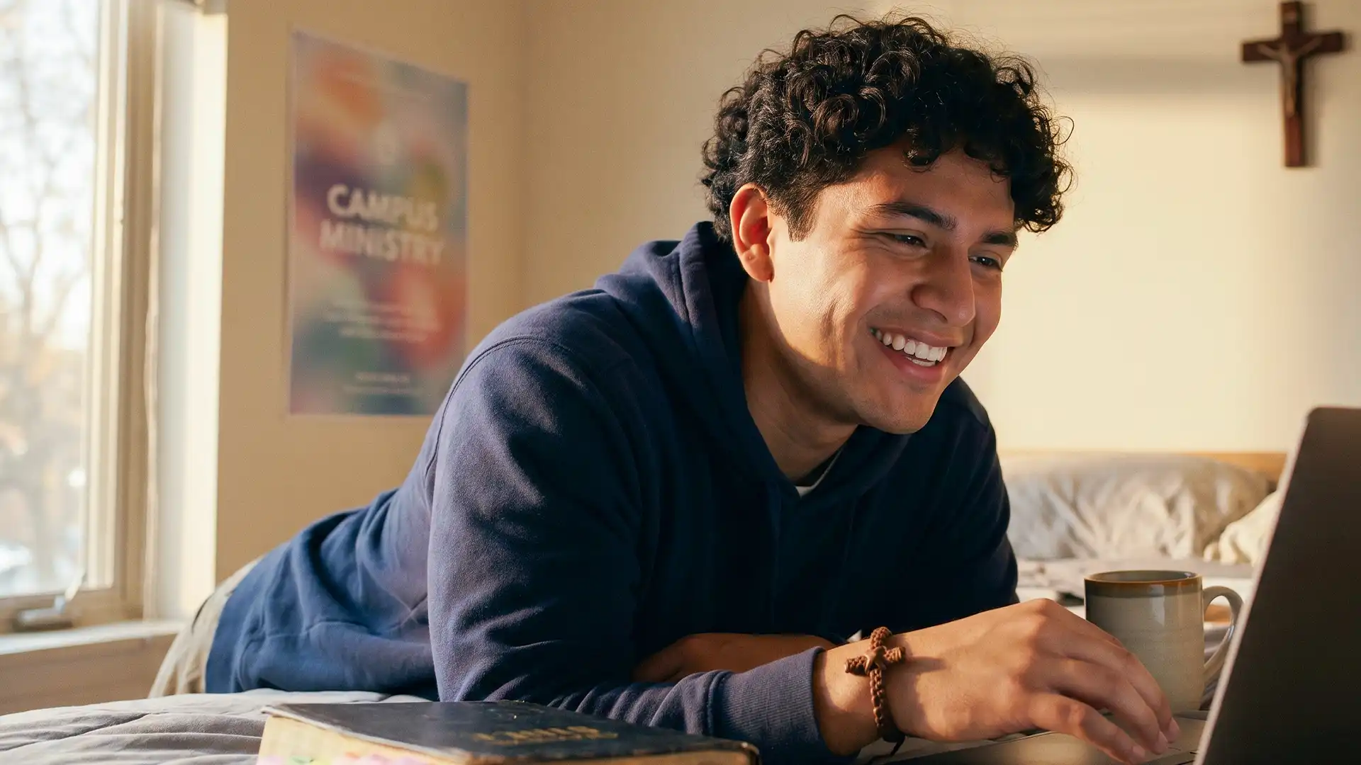 A smiling young man lying on his bed, engaged in an online faith-based coaching session on his laptop, with a Bible and campus ministry poster nearby.