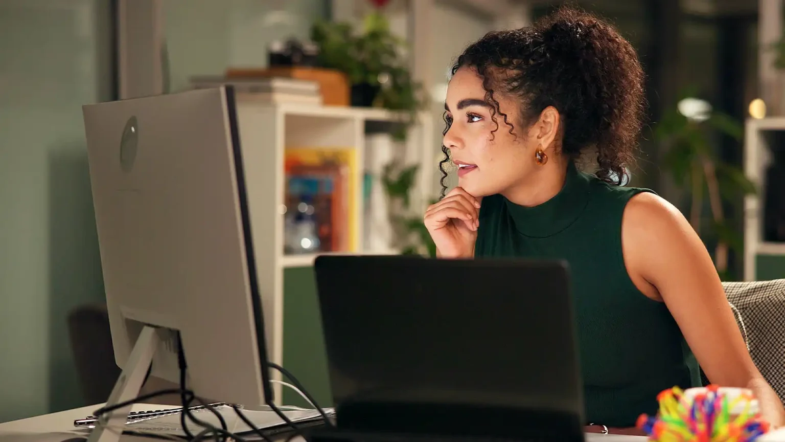 A focused woman with curly hair working late at a desk, contemplating personal growth on her computer in a softly lit home office with a bookshelf.
